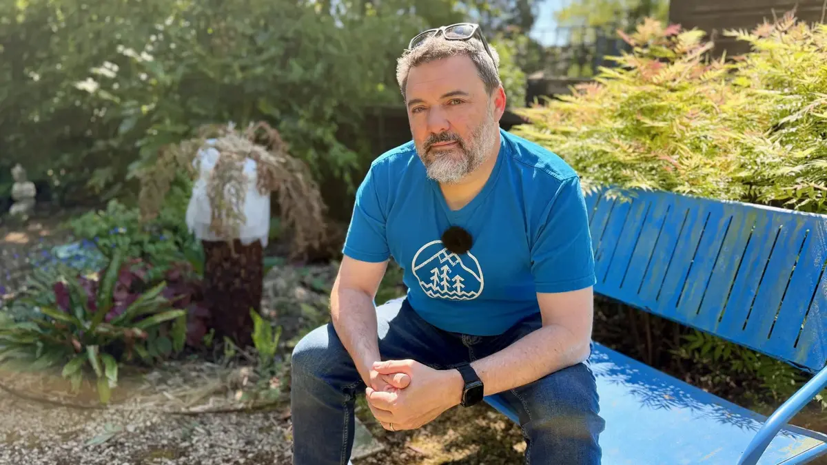 Chris Goor sitting on a blue bench in a garden, talking to camera