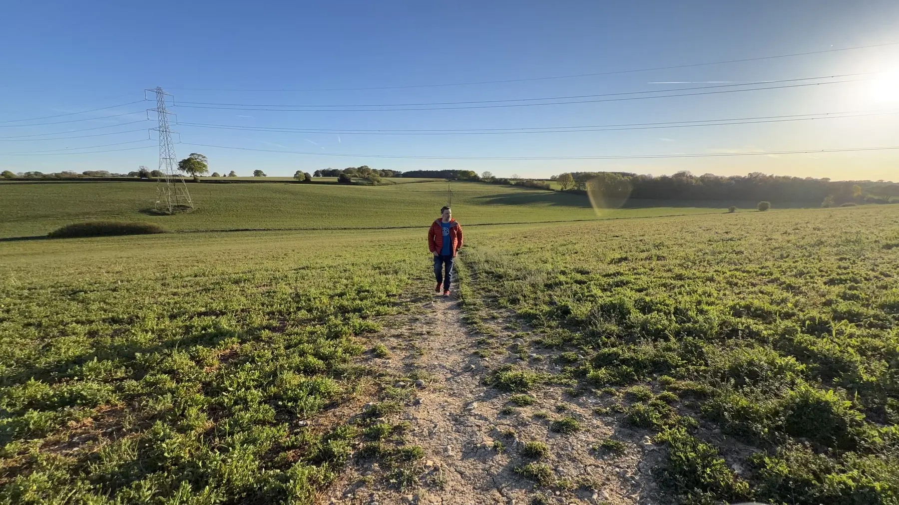 A person walking a dirt path towards camera across an open green field, pylon to the left, low sun flaring top right